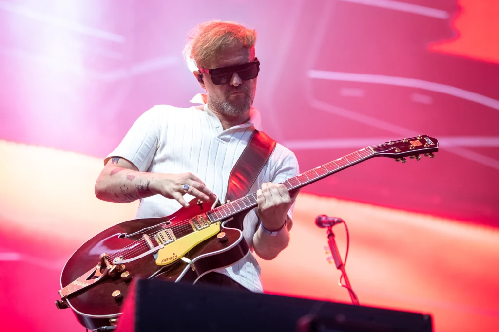 Organek podczas koncertu Mystic.30 Mężczyzna w okularach przeciwsłonecznych grający na gitarze elektrycznej na scenie, oświetlony kolorowym światłem podczas występu muzycznego.