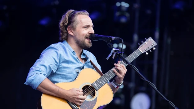 Muzyk w jasnej koszuli grający na gitarze akustycznej i śpiewający do mikrofonu podczas koncertu na scenie.