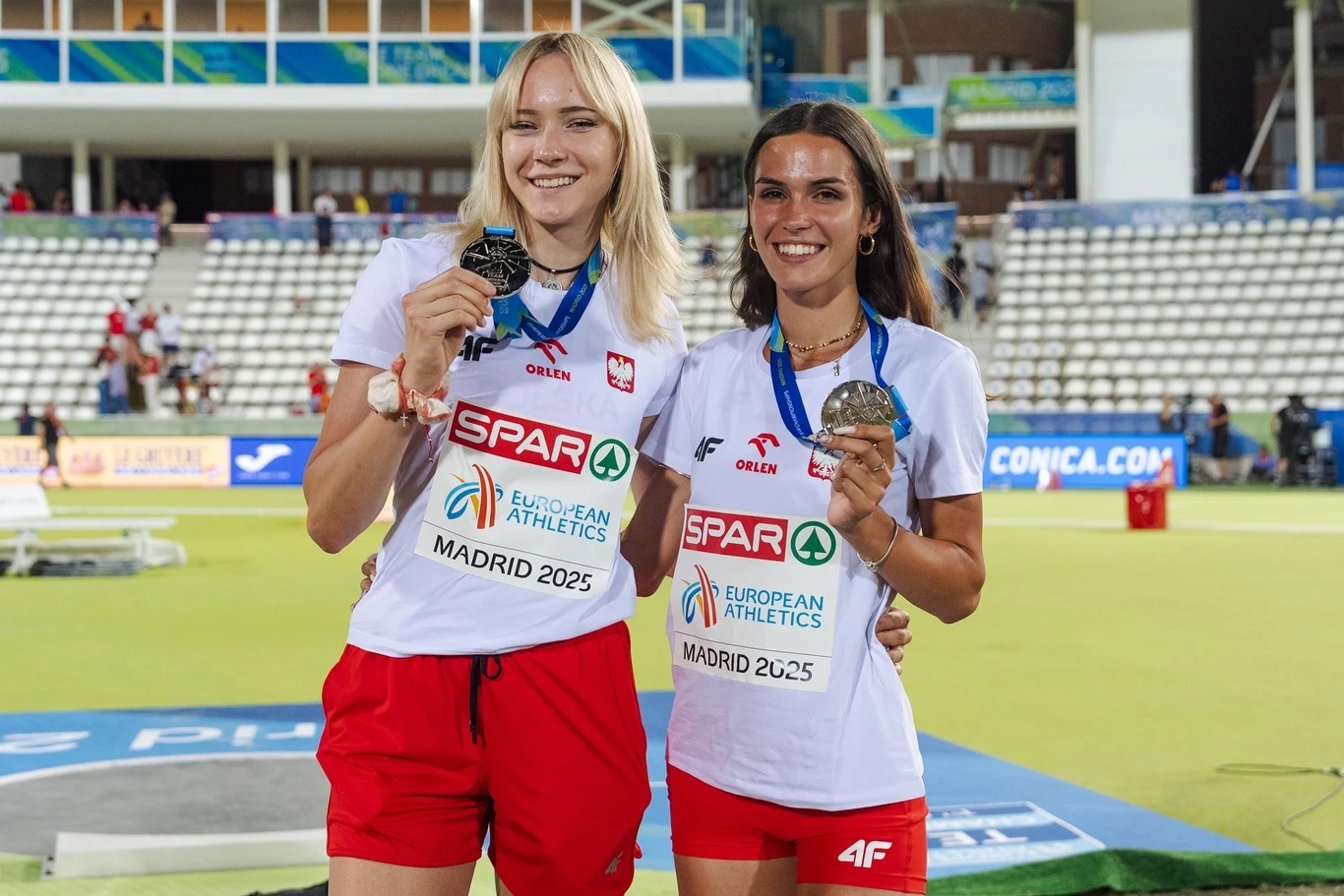Dwie młode sportsmenki w biało-czerwonych strojach reprezentacyjnych trzymają srebrne medale i pozują na stadionie lekkoatletycznym, na tle trybun i banerów reklamowych zawodów.