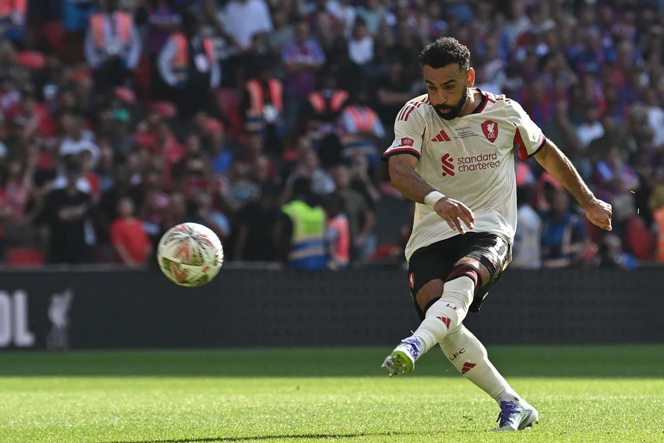 Piłkarz w białej koszulce Liverpoolu wykonuje kopnięcie piłki na zielonej murawie stadionu, w tle widać kibiców na trybunach.