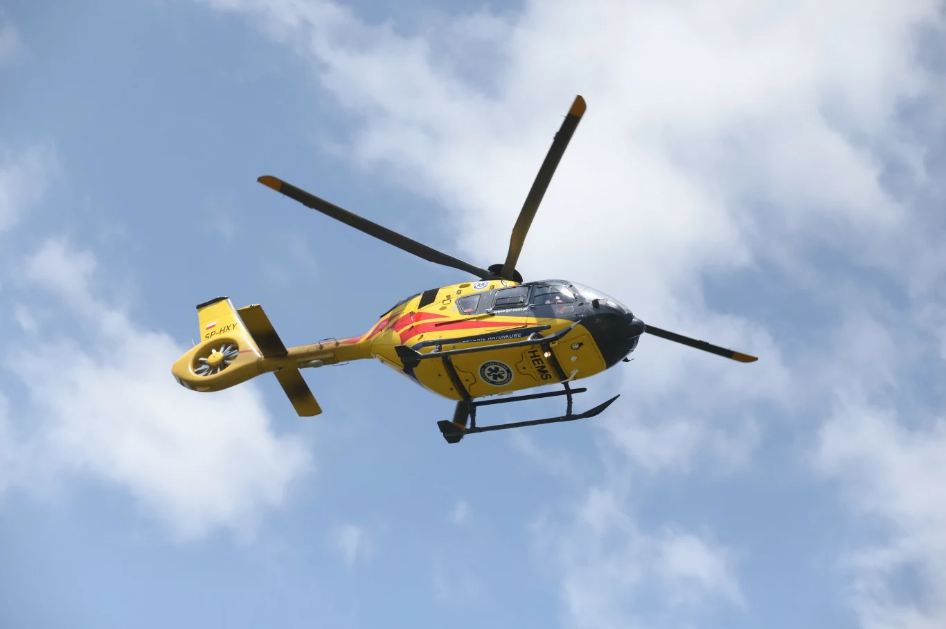 Pijany kierowca wjechał w grupę pielgrzymów. Są poszkodowani, wezwano śmigłowce LPR, zdj. ilustracyjne Żółto-czerwony helikopter ratunkowy leci na niebie z widocznym symbolem medycznym na burcie, na tle niebieskiego nieba z białymi chmurami.