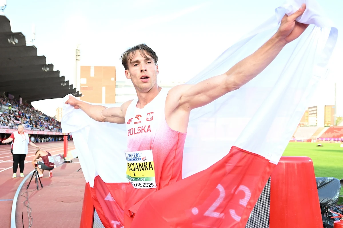 Hubert Trościanka Lekkoatleta w koszulce z napisem 'Polska', owinięty biało-czerwoną flagą narodową, świętuje sukces na stadionie lekkoatletycznym w obecności kibiców na trybunach.