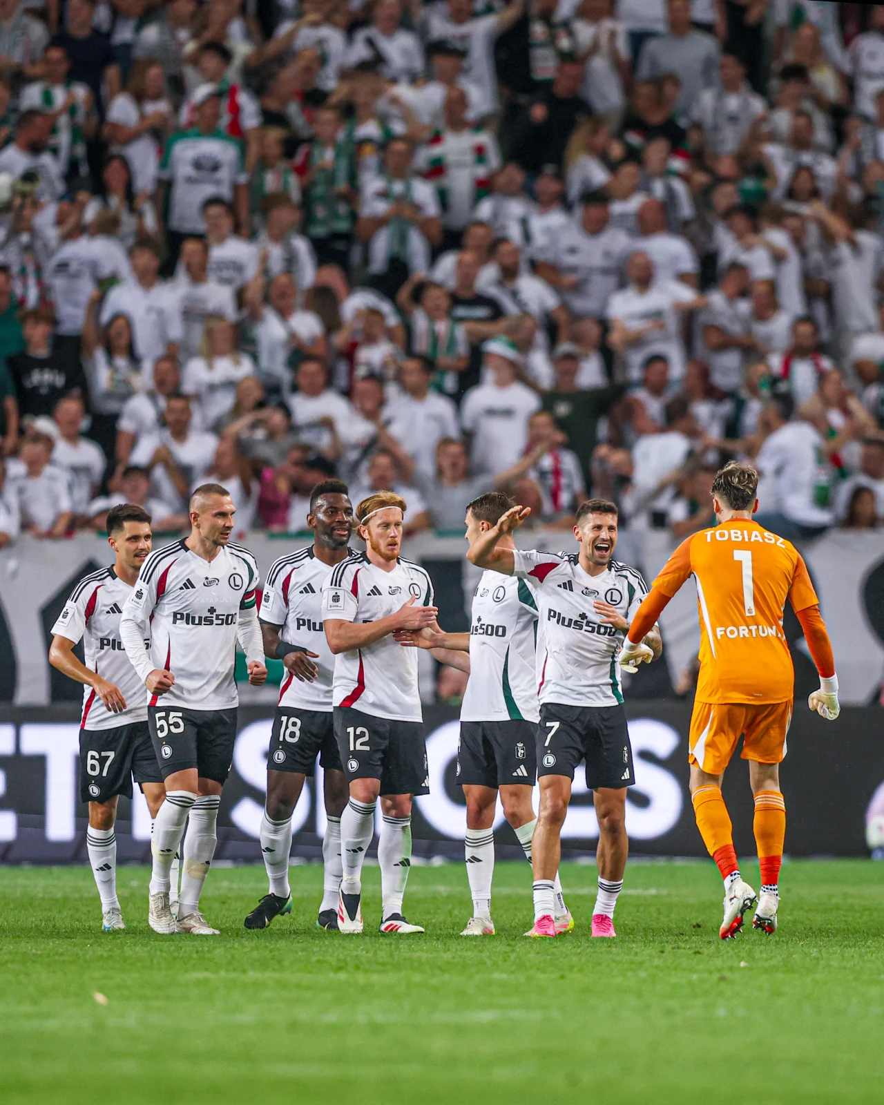 Grupa piłkarzy drużyny Legia Warszawa ubranych w białe stroje z logo sponsora świętuje na murawie z bramkarzem w pomarańczowym stroju, na trybunach tłum kibiców ubranych na biało.