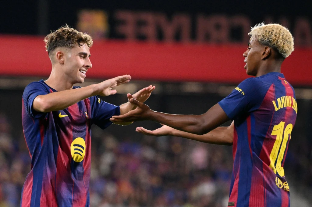 Lamine Yamal, Fermin Lopez Dwóch młodych piłkarzy drużyny FC Barcelona celebruje udaną akcję na boisku, ubrani w oficjalne stroje meczowe, w tle rozmazana publiczność na stadionie.