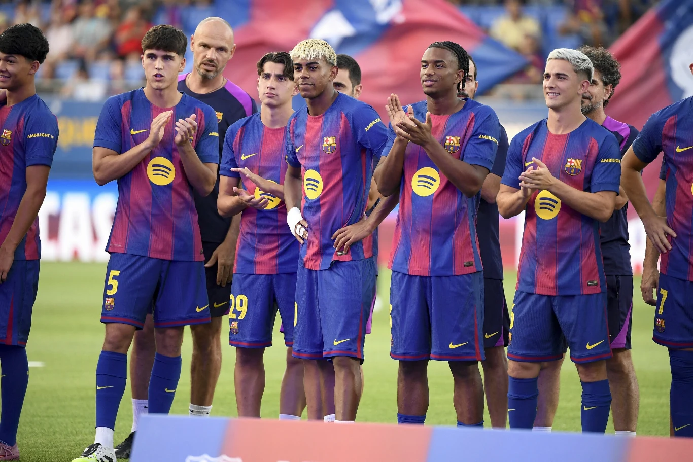 Grupa młodych piłkarzy drużyny FC Barcelona w niebiesko-czerwonych strojach stoi razem na murawie stadionu, kilku z nich klaska, w tle rozmyte flagi i kibice