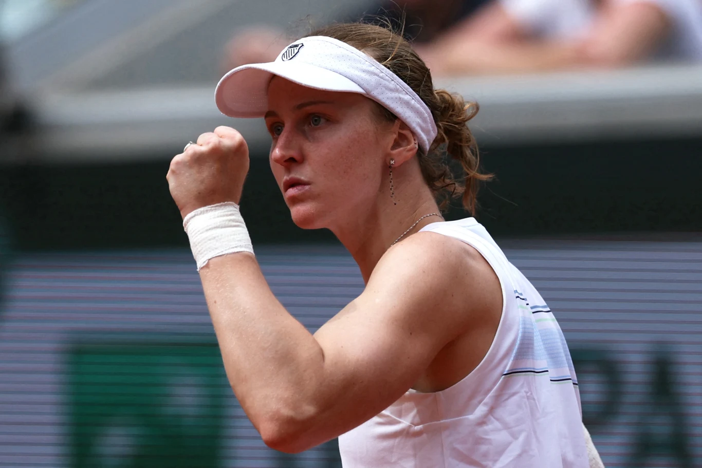 Tenisistka w białym stroju i białej czapce z daszkiem, z opaską na nadgarstku, zaciska pięść w geście triumfu podczas meczu sportowego, wyrażając determinację i koncentrację.