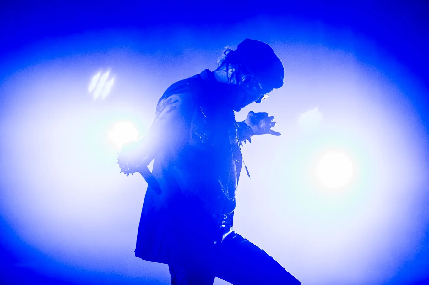 Atilla Csihar (Mayhem) Sylwetka osoby trzymającej mikrofon na tle intensywnie niebieskiego światła scenicznego, dynamiczna poza sugerująca występ muzyczny.