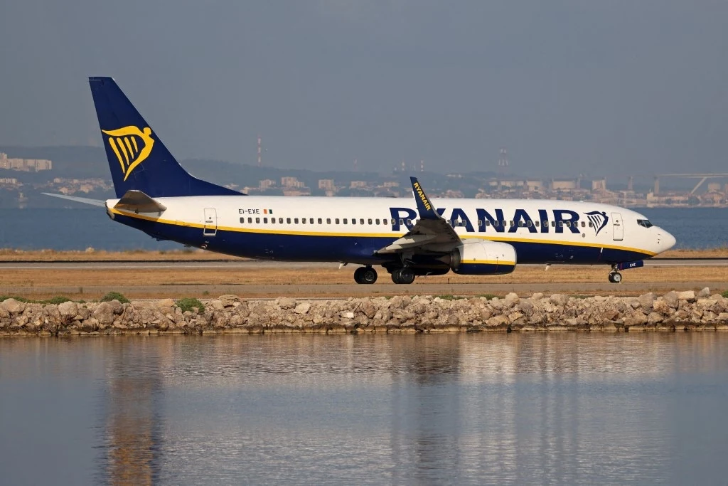 Pilot samolotu linii Ryanair zemdlał podczas lotu (zdjęcie ilustracyjne) Samolot pasażerski linii Ryanair porusza się po pasie startowym w pobliżu wody, w tle widoczna zabudowa przemysłowa lub portowa.