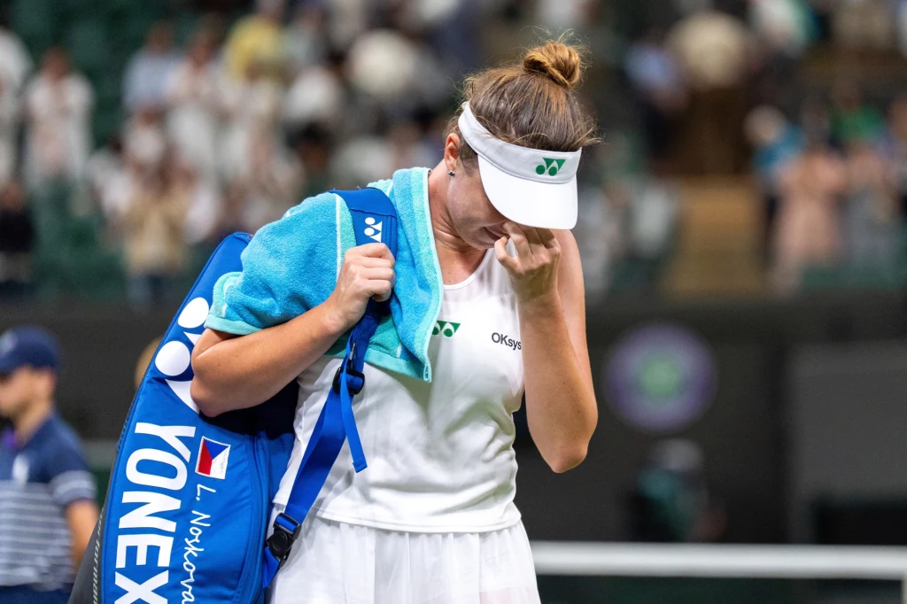 Zawodniczka tenisowa w białym stroju opuszcza kort z ręcznikiem na ramieniu i torbą sportową na plecach, wyraźnie zasmucona, ociera oczy, prawdopodobnie po przegranym meczu