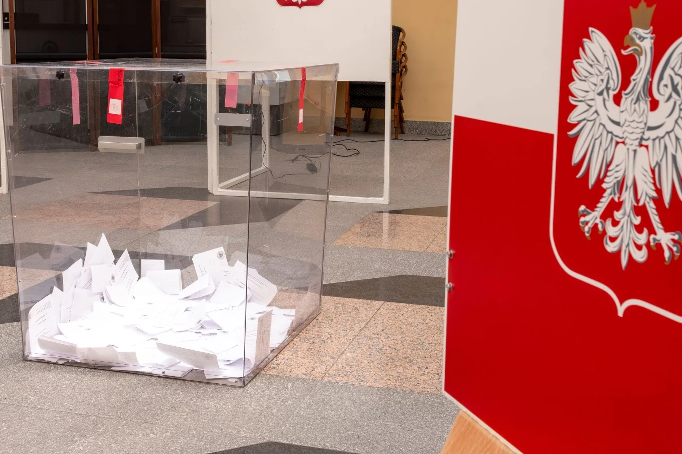 Wyniki I tury wyborów prezydenckich w Zabrzu. Zdjęcie ilustracyjne Przezroczysta urna wypełniona kartami do głosowania, w tle kabiny wyborcze z widocznym godłem Polski oraz fragment sali wyborczej.