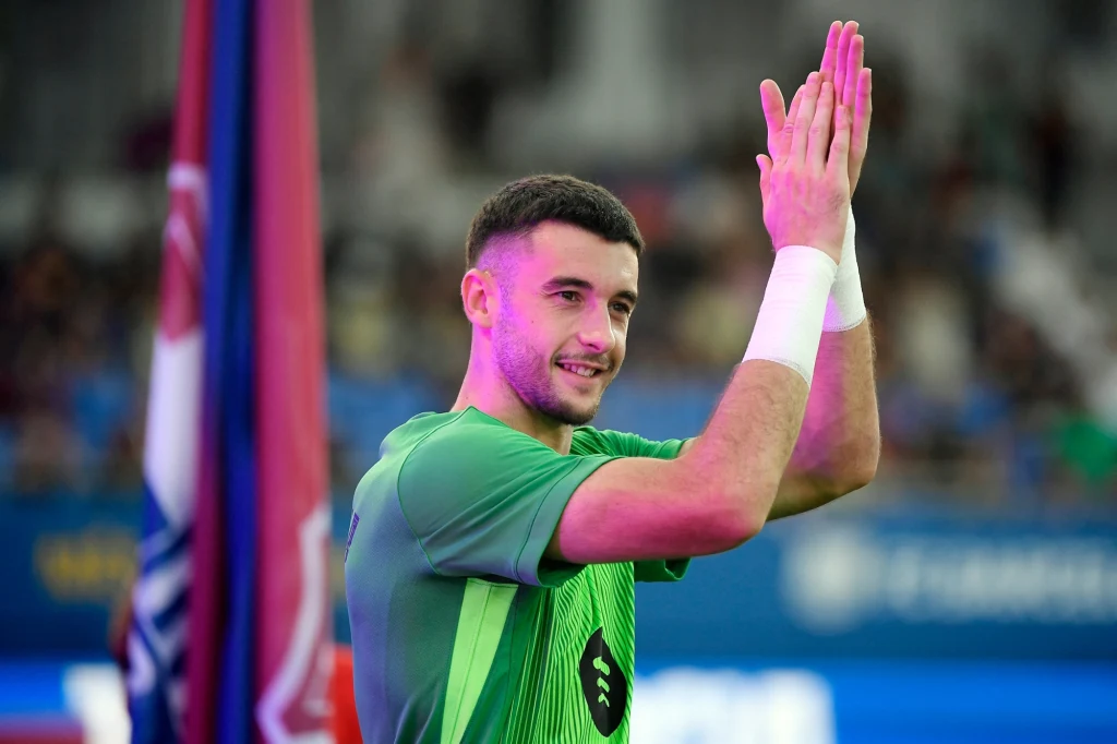 Młody mężczyzna w zielonej sportowej koszulce bije brawo i uśmiecha się, stojąc na tle flagi i rozmazanego stadionu z widownią.