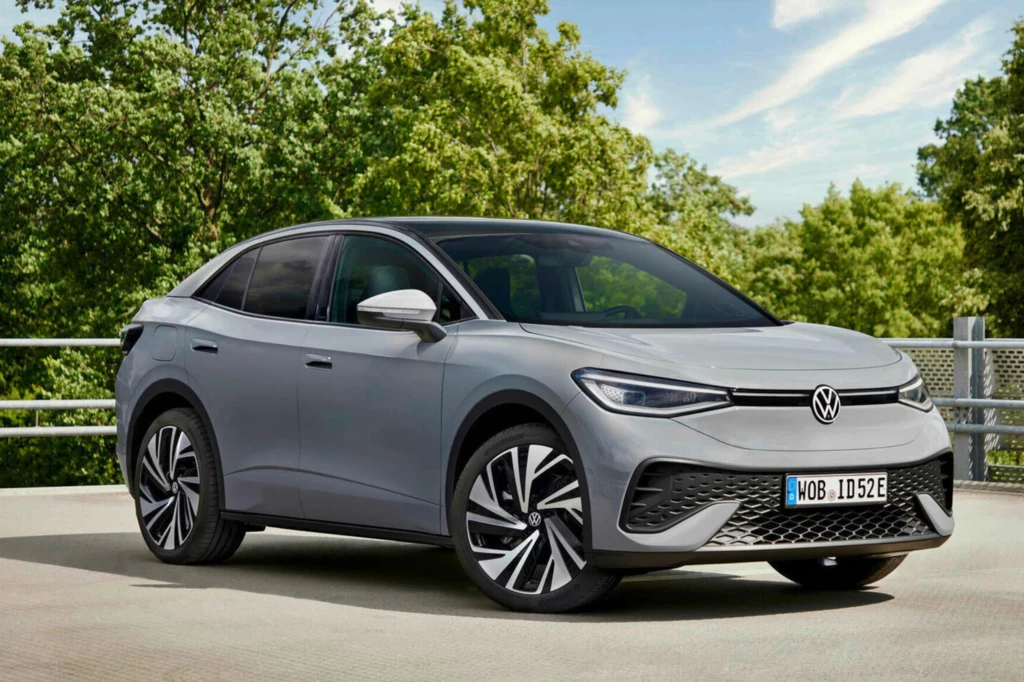 Srebrny samochód elektryczny Volkswagen ID.5 stoi zaparkowany na tle zielonych drzew, na otwartym terenie z betonowym podłożem, widoczne są charakterystyczne futurystyczne felgi i nowoczesny design pojazdu.