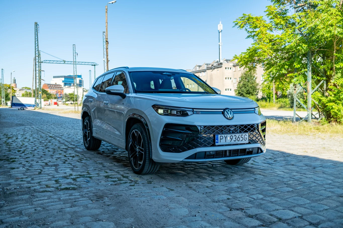 Z ceną na poziomie 250 tys. zł Tayron wchodzi już na teren marek premium - za taką kwotę można myśleć o zakupie choćby Audi Q5. Nowoczesny biały samochód marki Volkswagen stoi zaparkowany na brukowanej ulicy, otoczony zielenią i zabudową miejską. Numery rejestracyjne pojazdu widoczne na przodzie. Tło stanowią słupy trakcyjne i budynki mieszkalne.