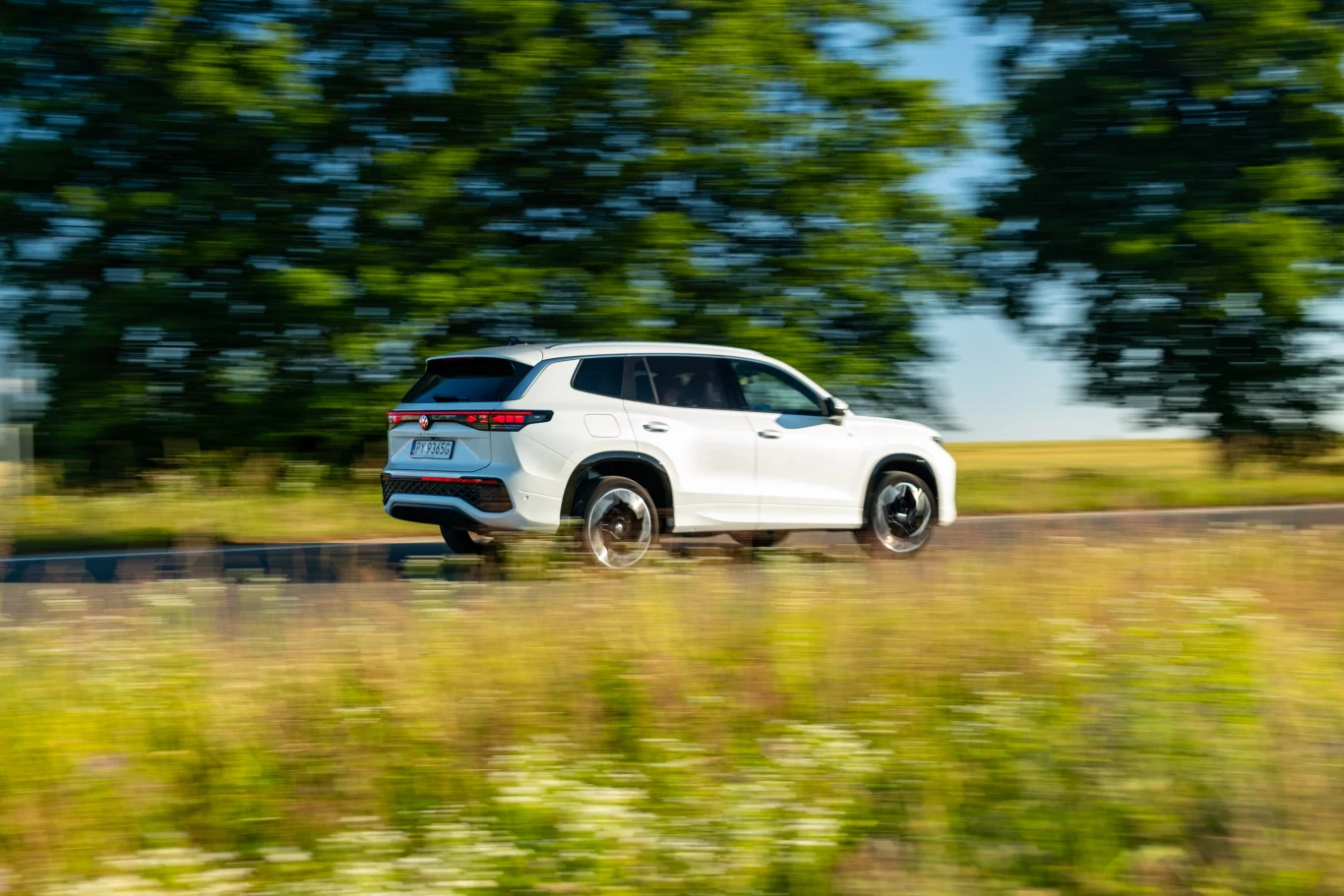 Auto prowadzi się neutralnie i przewidywalnie. Duże koła z oponami o zdrowym profilu przyczyniają się do wysokiego komfortu jazdy. Biały SUV jedzie z dużą prędkością po asfaltowej drodze, w tle rozmazane drzewa i zielone pole, wokół samochodu rozmyta trawa tworząca efekt dynamicznego ruchu.