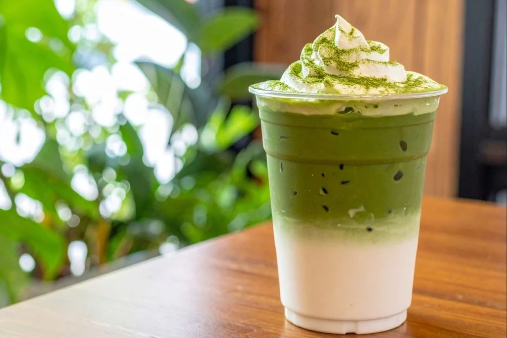 Matcha świetnie skomponuje się z mlekiem, a nawet lodami stanowiąc pyszny i zdrowy deser Plastikowy kubek z warstwowym napojem składającym się z białego mleka i zielonej herbaty matcha z lodem oraz bitą śmietaną i posypką z zielonej herbaty, ustawiony na drewnianym stole, w tle zielone liście roślin.