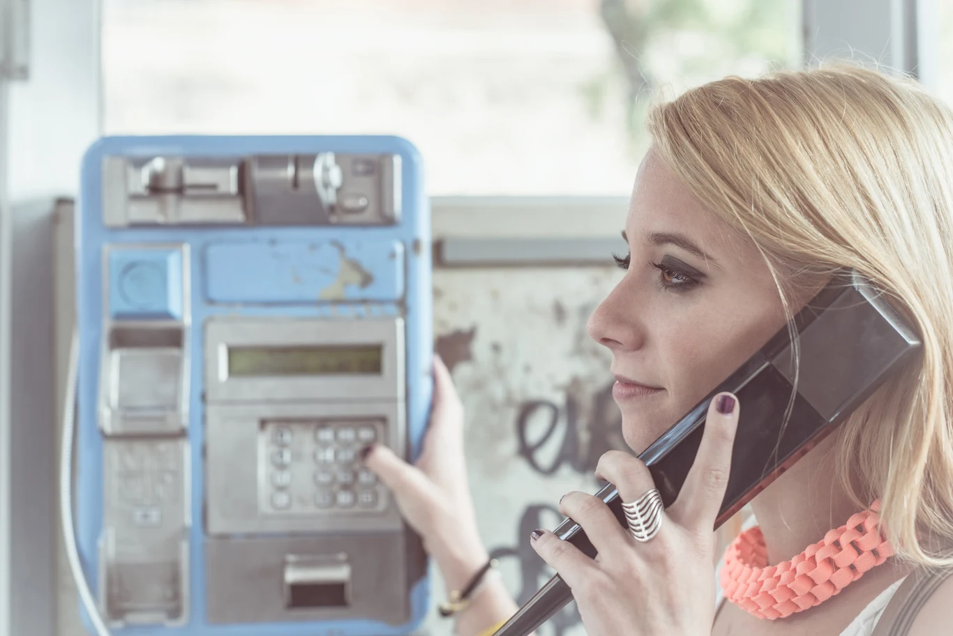 Wraz z wieloma urządzeniami sprzed lat w zapomnienie odeszły charakterystyczne dźwięki. Czy jeszcze je pamiętasz? Młoda kobieta rozmawia przez telefon publiczny, trzymając słuchawkę przy uchu i patrząc w bok, w tle widać niebieski automat telefoniczny.
