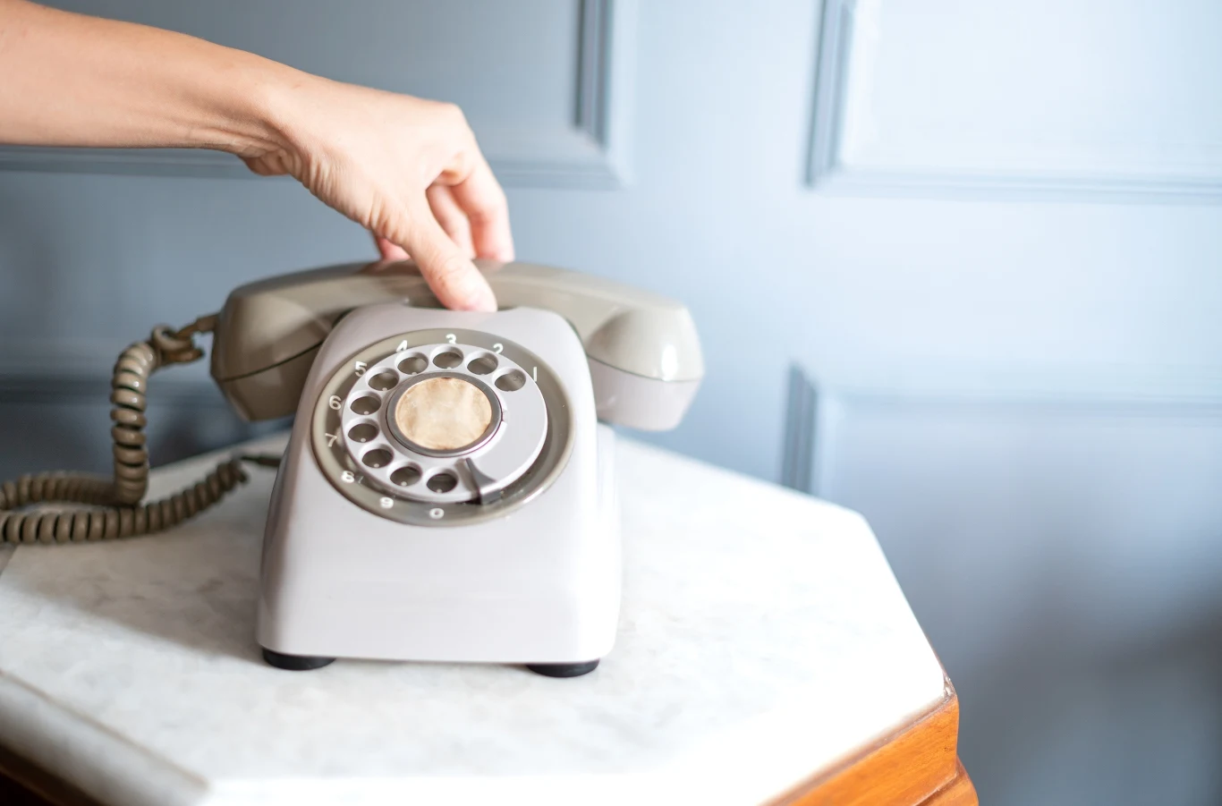 Telefony stacjonarne z tarczą wydawały charakterystyczny dźwięk przy wybieraniu kolejnych cyfr Ręka sięgająca po szary, klasyczny telefon stacjonarny z tarczą obrotową, ustawiony na blacie z marmuru przy jasnoniebieskiej ścianie.