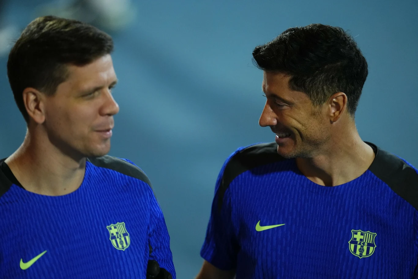 Dwóch mężczyzn ubranych w niebieskie koszulki sportowe z herbem klubu piłkarskiego i żółtym logo Nike rozmawia ze sobą i uśmiecha się, stojąc obok siebie na tle jasnego, rozmytego tła.