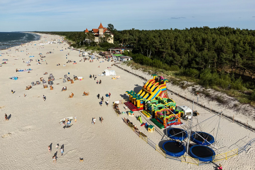 Łeba to z jednej strony rozbudowana infrastruktura turystyczna, a z drugiej miejsce, w którym da się znaleźć bardziej zaciszne zakątki Szeroka piaszczysta plaża nad morzem, na pierwszym planie kolorowy plac zabaw z dmuchanymi zjeżdżalniami i trampolinami, w tle las sosnowy oraz charakterystyczny budynek z czerwonym dachem, rozproszeni spacerowicze korzystający z wypoczynku.