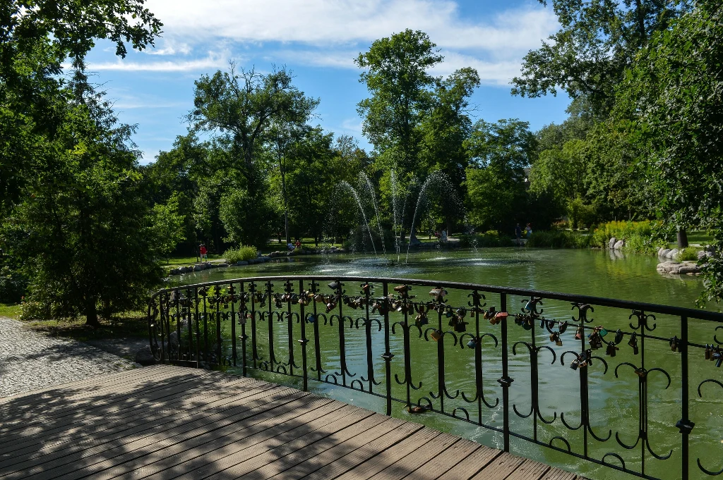 Park Zdrojowy w Ciechocinku Mostek ozdobiony metalową balustradą nad stawem, z fontanną na środku wody, otoczony zielenią drzew i błękitnym niebem w tle.