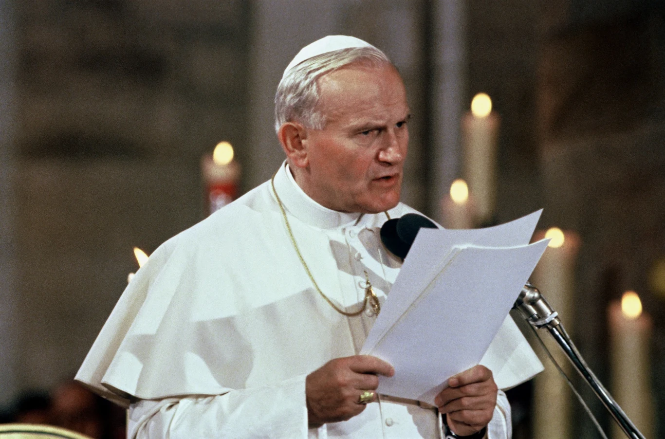 Papieska postać w białych szatach podczas przemówienia, trzymająca kartki papieru przy mikrofonie, w tle widoczne zapalone świece.