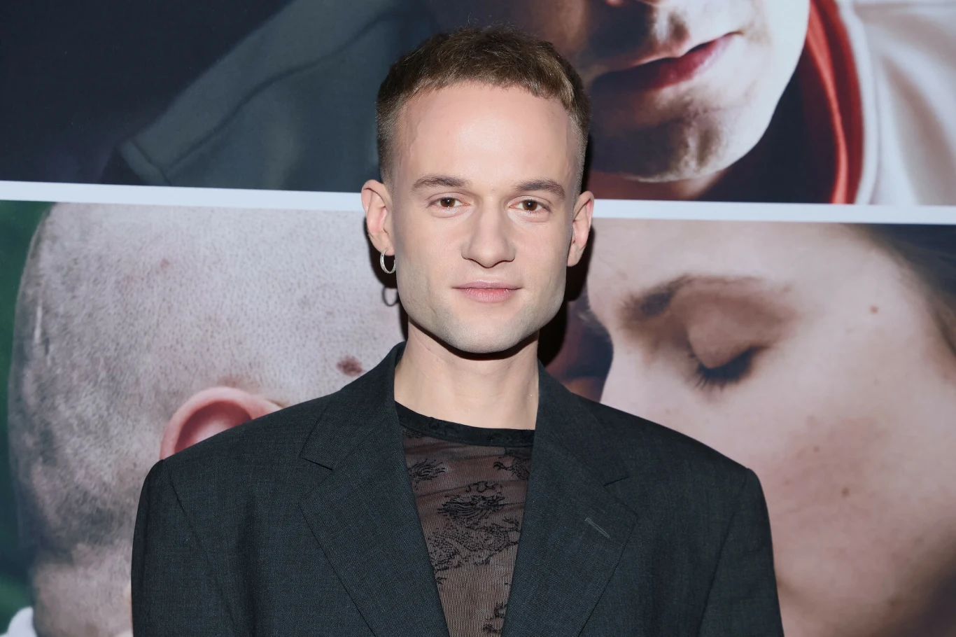 Kamil Studnicki będzie jednym z uczestników "Twoja Twarz Brzmi Znajomo" Uśmiechnięty mężczyzna w ciemnej marynarce stoi na tle plakatu filmowego z dużymi twarzami innych osób.