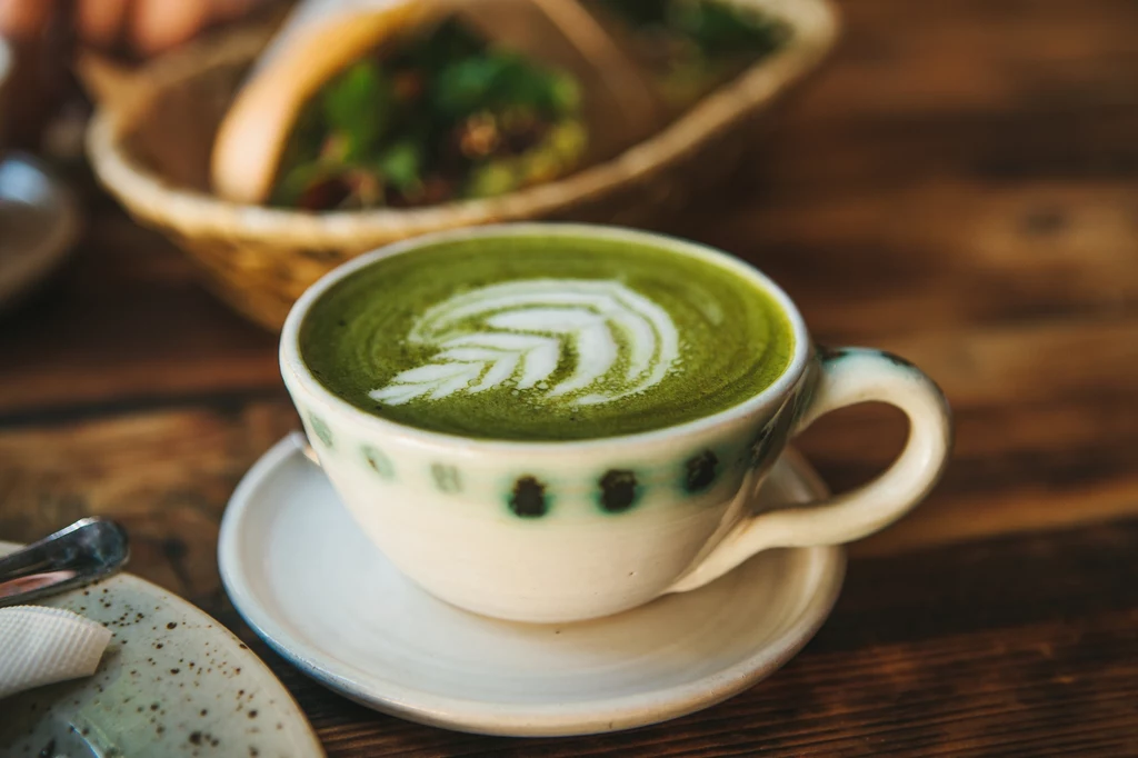 Kubek z zieloną herbatą matcha z artystycznie wykonaną pianką na wierzchu stoi na drewnianym stole, w tle widoczny koszyk z jedzeniem.