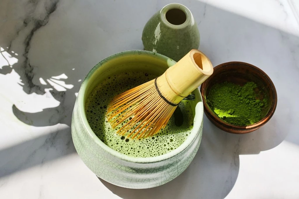 Piękna zielona miska z ubijaną herbatą matcha, bambusowa miotełka do mieszania, mała miseczka z proszkiem matcha oraz ceramiczna karafka ustawione na jasnym tle, w świetle dziennym padającym przez okno.