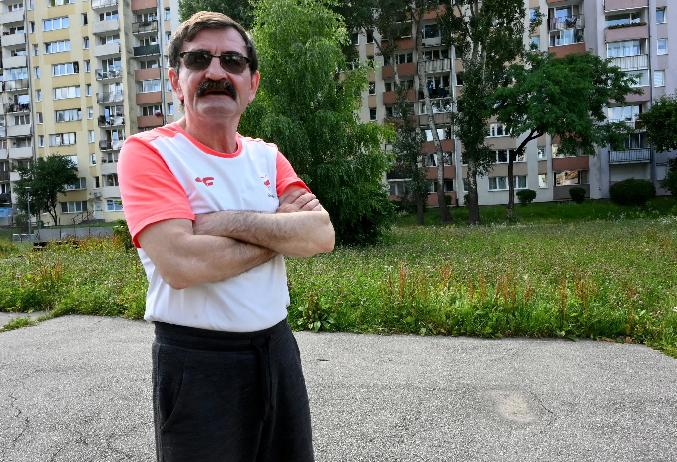 Mężczyzna w okularach przeciwsłonecznych i białej koszulce z czerwonymi rękawami stoi z założonymi rękami na tle bloków mieszkalnych oraz zarośniętego trawnika.