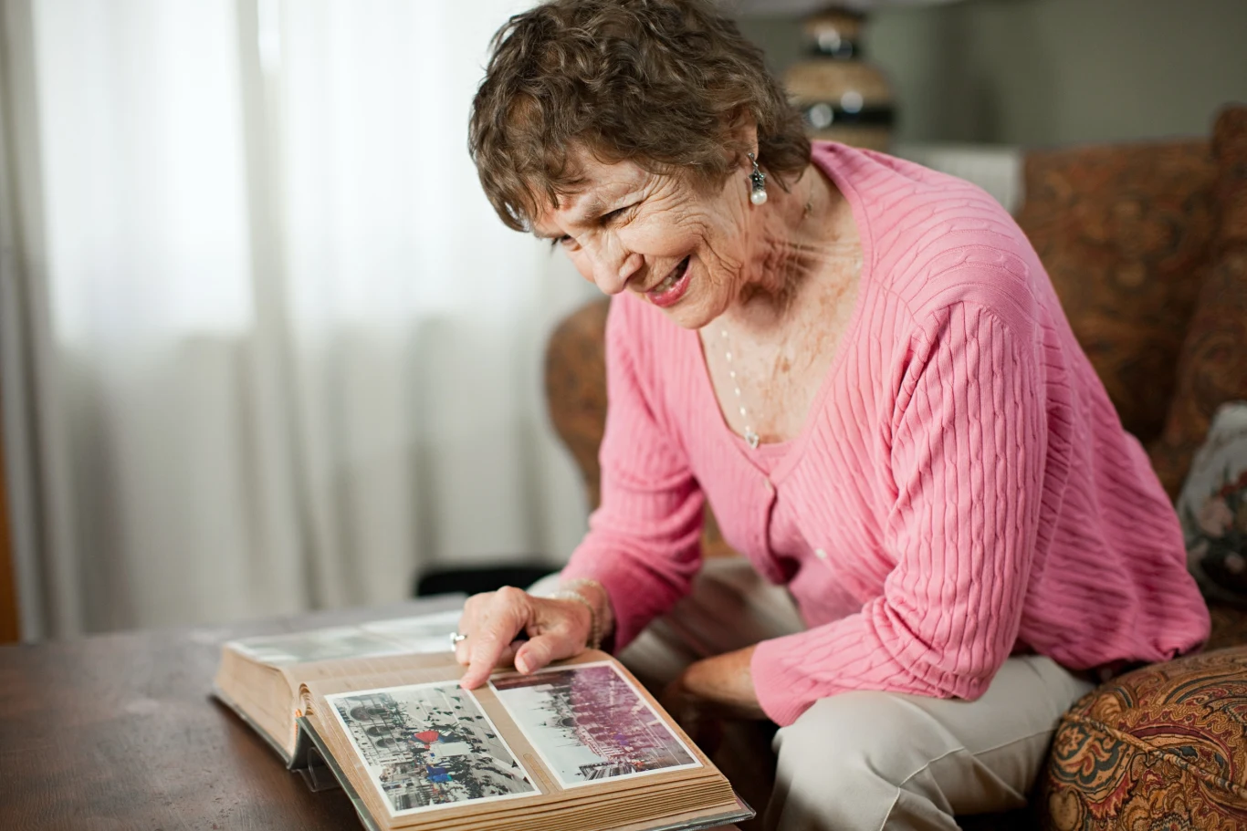 Starsza kobieta w różowym swetrze siedzi na kanapie i z uśmiechem przegląda album ze starymi zdjęciami, zanurzona w nostalgii.