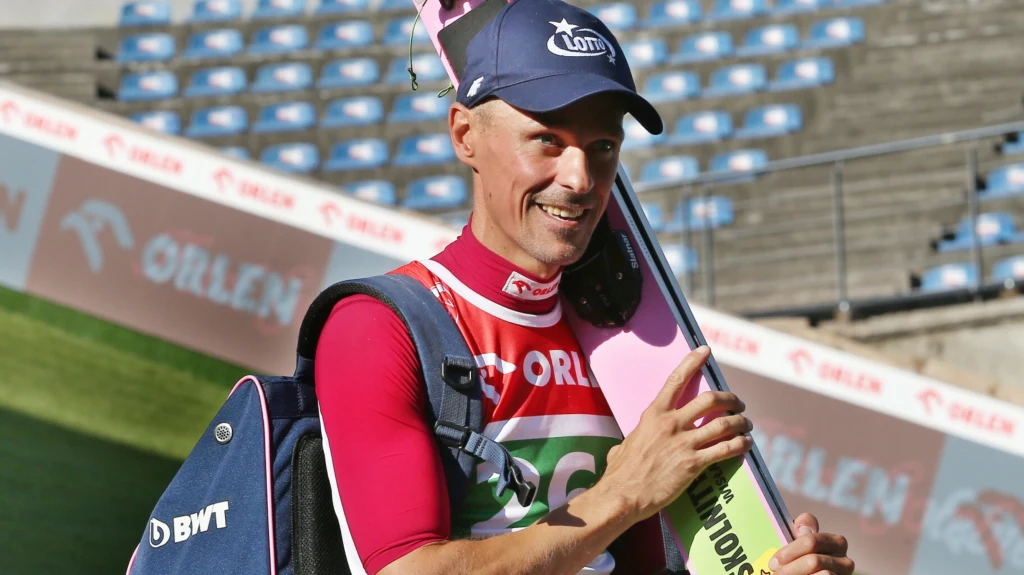 Piotr Żyła Uśmiechnięty skoczek narciarski w stroju sportowym i czapce z logotypem, trzyma narty na ramieniu oraz torbę sportową, w tle puste trybuny stadionu sportowego.