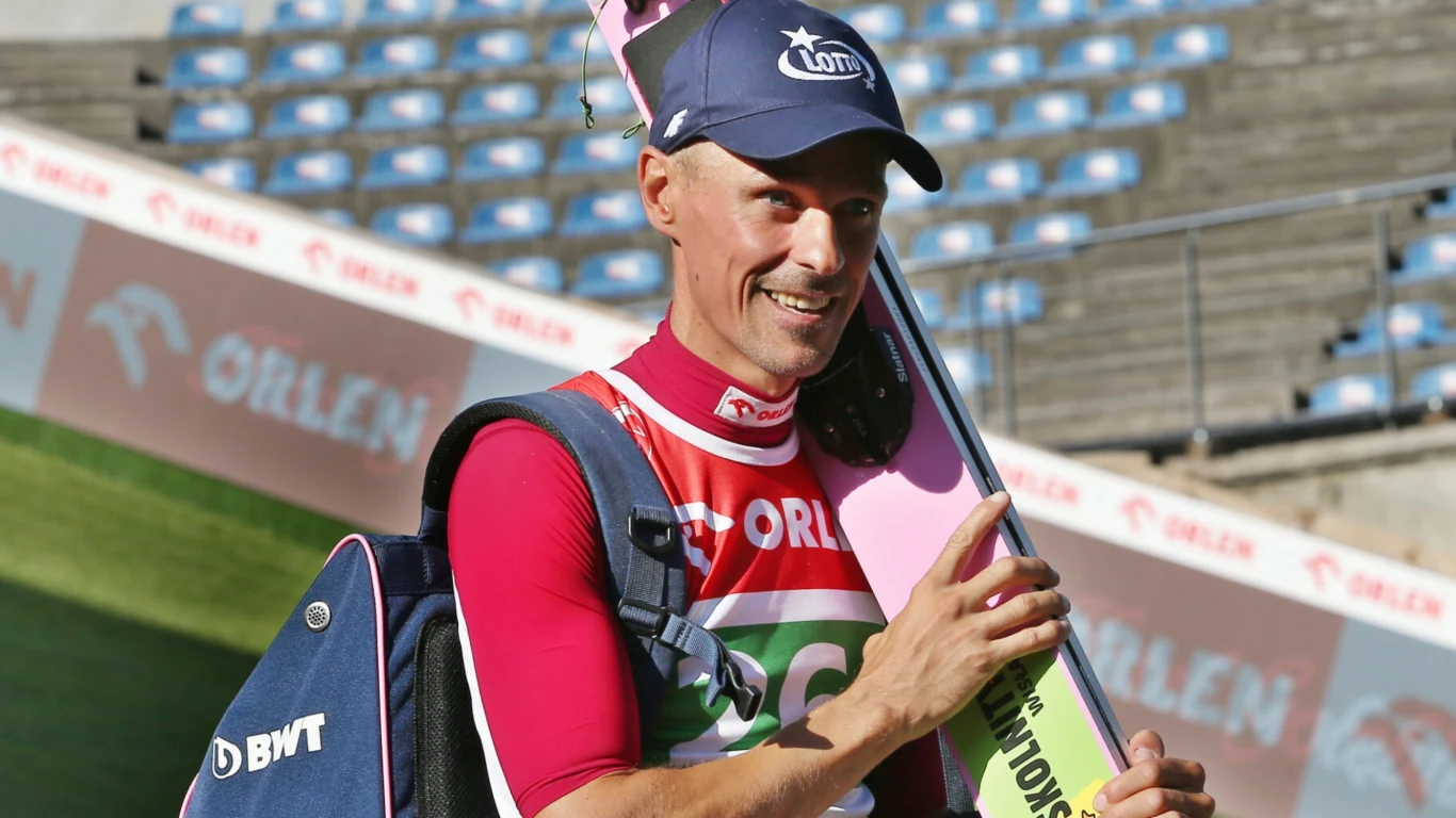 Uśmiechnięty skoczek narciarski w stroju sportowym i czapce z logotypem, trzyma narty na ramieniu oraz torbę sportową, w tle puste trybuny stadionu sportowego.