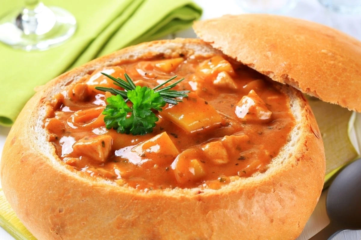 Gulasz podany w wydrążonym bochenku chleba, dekorowany świeżą natką pietruszki i gałązką rozmarynu, obok przykrywka z chleba na jasnozielonej serwetce.