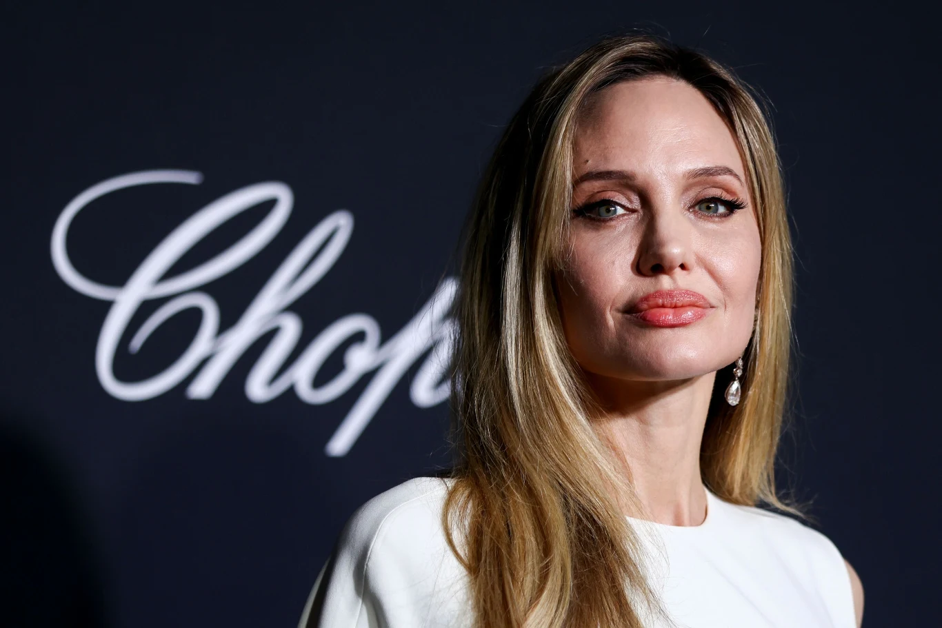 Angelina Jolie w eleganckiej białej sukni z długimi blond włosami pozuje na tle ciemnej ściany z białym napisem Chopard.