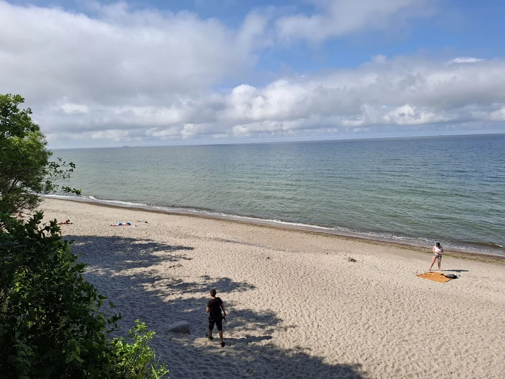 Piaszczysta plaża nad morzem z dwoma osobami, jedna z nich chodzi po piasku, a druga przygotowuje się do wejścia do wody; w tle spokojna tafla wody i niebo z rozproszonymi chmurami.