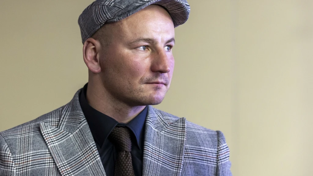 Mężczyzna w marynarce w kratę, czarnej koszuli, brązowym krawacie i kaszkiecie patrzący w bok na jasnym, jednolitym tle.