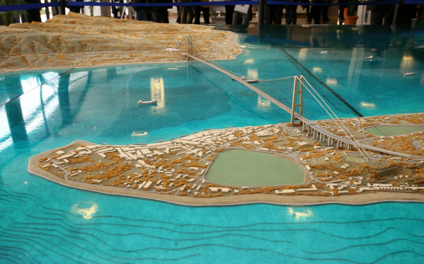 Model Mostu Mesyńskiego, który połączy Kalabrię z Sycylią Makieta mostu wiszącego łączącego dwa fragmenty lądu oddzielonego wodą, szczegółowo odwzorowane zabudowania miejskie i tereny zielone w otoczeniu mostu.