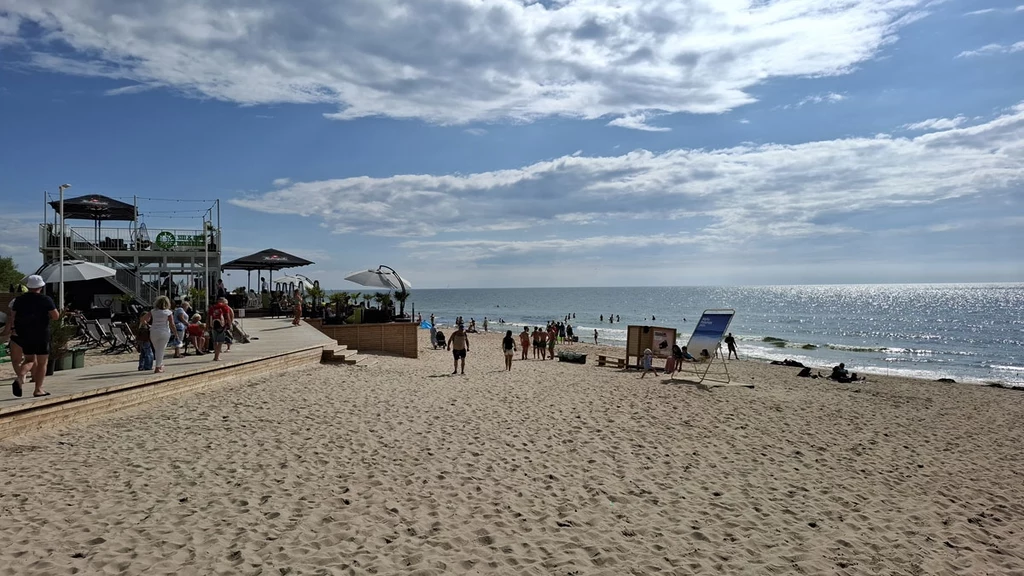 Szeroka, piaszczysta plaża z ludźmi spacerującymi i odpoczywającymi przy brzegu morza, wejście na plażę z budynkiem gastronomicznym i strefą wypoczynkową, spokojne fale oraz częściowo zachmurzone niebo.