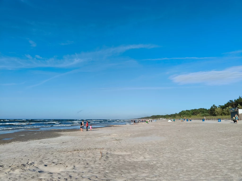 Zdjęcie zrobione w sierpniowe popołudnie. Wystarczy przejść kawałek dalej od głównych wejść, aby cieszyć się spokojem Szeroka, piaszczysta plaża z kilkoma spacerującymi osobami, spokojne fale morskie oraz pas zieleni z drzewami po prawej stronie i błękitne, niemal bezchmurne niebo.
