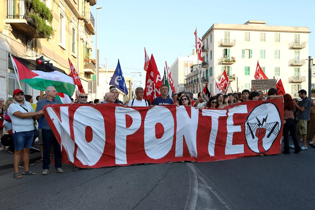 Grupa ludzi idąca ulicą z dużą czerwoną flagą z napisem NO PONTE, w tle widać flagi różnych organizacji oraz miejską zabudowę.