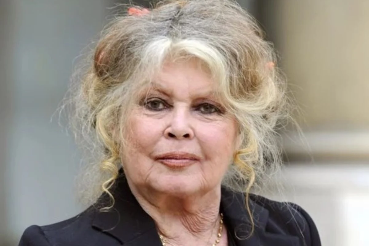  Brigitte Bardot o jasnych, rozwianych włosach z lekkimi lokami, ubrana w ciemny żakiet, patrzy prosto w obiektyw. Jej twarz wyrażająca spokój i powagę, na tle rozmytego wnętrza.