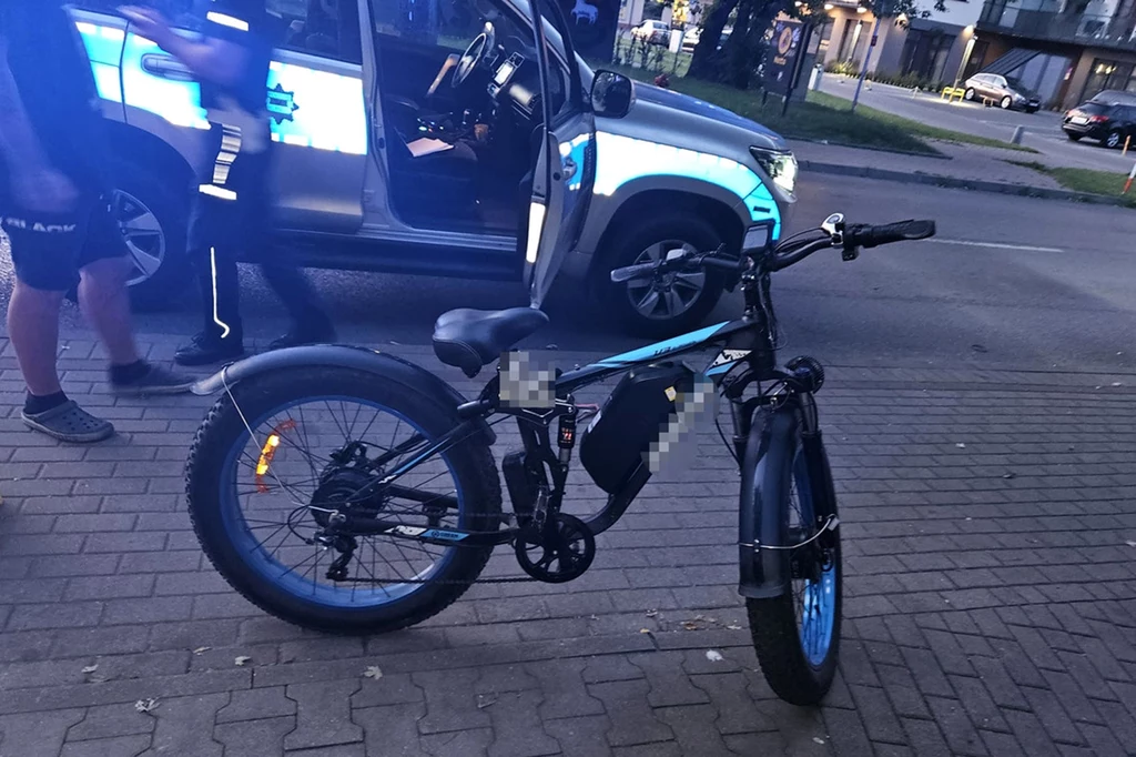 Elektryczny rower z szerokimi oponami stoi na chodniku, obok zaparkowany samochód policyjny z otwartymi drzwiami, widoczne postaci w sportowych ubraniach, fragment miejskiego krajobrazu z ulicą i zabudowaniami w tle