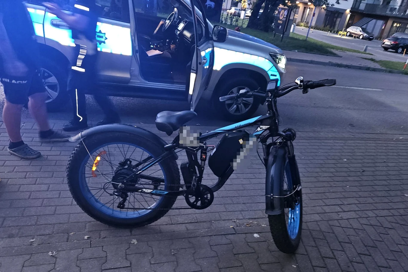 Elektryczny rower z szerokimi oponami stoi na chodniku, obok zaparkowany samochód policyjny z otwartymi drzwiami, widoczne postaci w sportowych ubraniach, fragment miejskiego krajobrazu z ulicą i zabudowaniami w tle