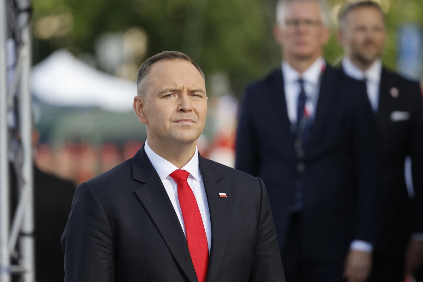 Prezydent RP Karol Nawrocki Mężczyzna w ciemnym garniturze z czerwoną krawatą i białą koszulą stoi na pierwszym planie, w tle dwaj inni mężczyźni w garniturach, tło rozmyte.