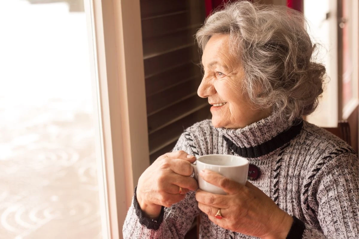 Starsza kobieta z siwymi włosami w ciepłym swetrze trzymająca filiżankę, patrzy przez okno z lekkim uśmiechem na twarzy, siedząc w przytulnym wnętrzu.