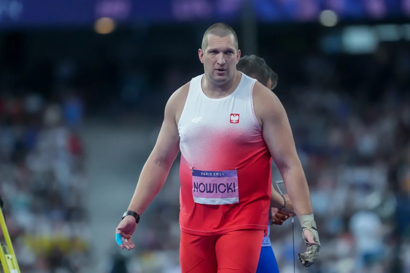 Lekkoatleta w stroju reprezentacji Polski podczas zawodów lekkoatletycznych na stadionie, z tabliczką z nazwiskiem Nowicki, w tle rozmazani kibice i inny sportowiec.