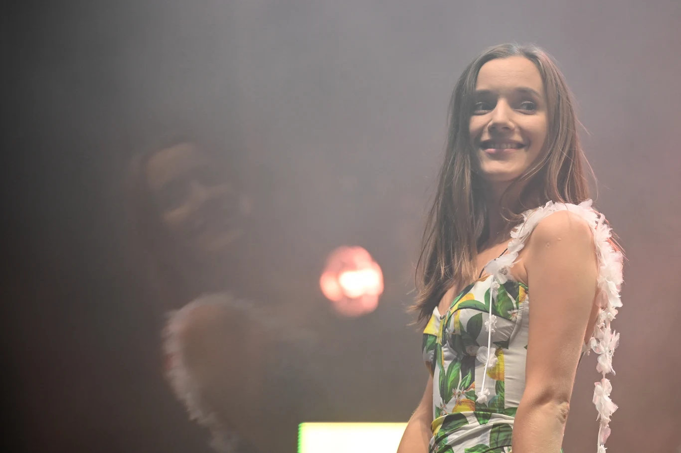 Utwory sanah i Skolima towarzyszą pielgrzymom! Uśmiechnięta kobieta w sukience z motywem roślinnym stoi na scenie, w tle widoczne są rozproszone światła i delikatny dym teatralny, obok kobiety odbicie światła w kształcie okręgu.