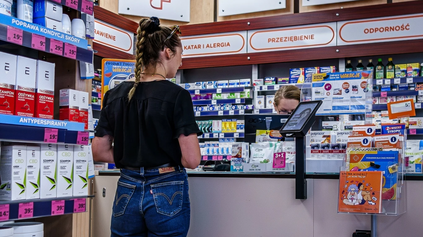 Aptekarze chcą być wyłączeni z systemu kaucynego Kobieta stoi przy ladzie w aptece, rozmawia z farmaceutką, za którą widoczne są półki wypełnione lekami, suplementami i innymi produktami zdrowotnymi, a na ścianach znajdują się tabliczki informujące o działach leków.