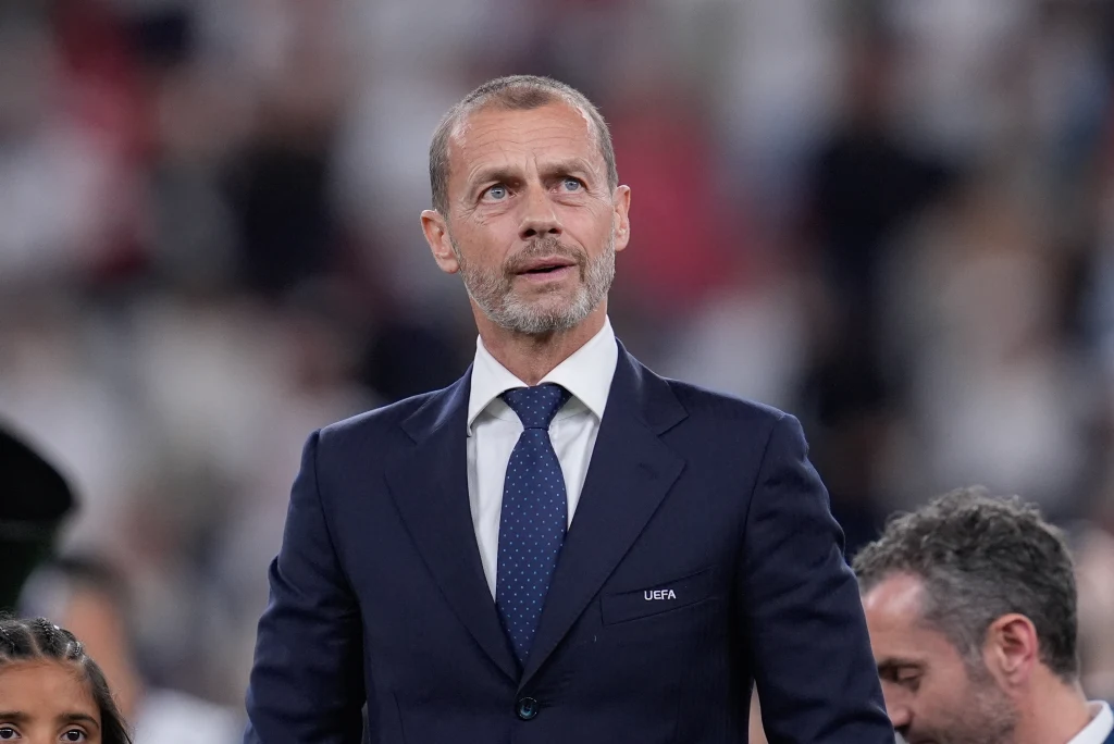 Mężczyzna w garniturze i krawacie stoi w tłumie, skupiony, na tle rozmytych postaci w jasnym otoczeniu.