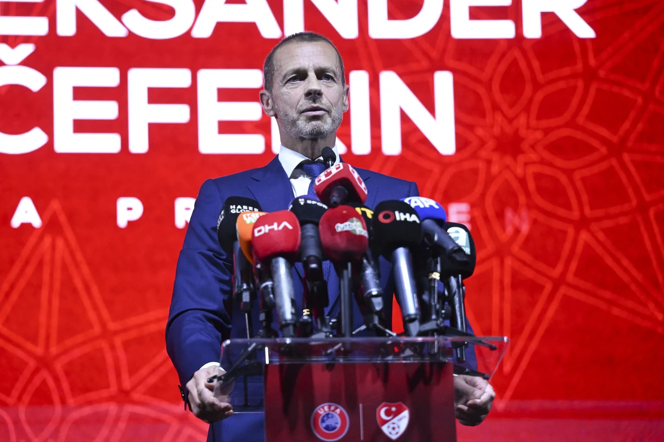 Aleksander Ceferin Mężczyzna w garniturze przemawia na tle czerwonej ściany z dużym napisem oraz wzorem, przed nim ustawionych jest wiele mikrofonów różnych mediów.